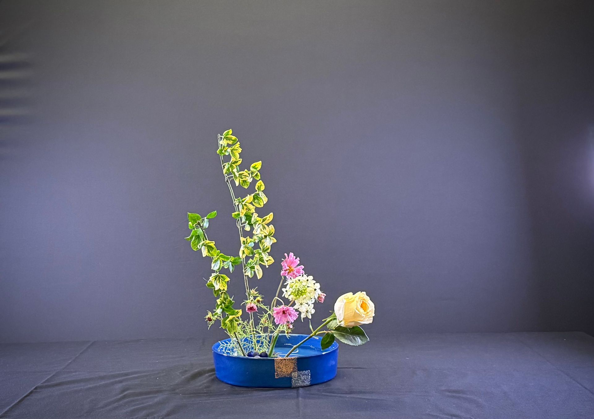 Bloemstuk in een blauwe vaas. Groene, gele, roze en witte bloemen tegen een grijze achtergrond.