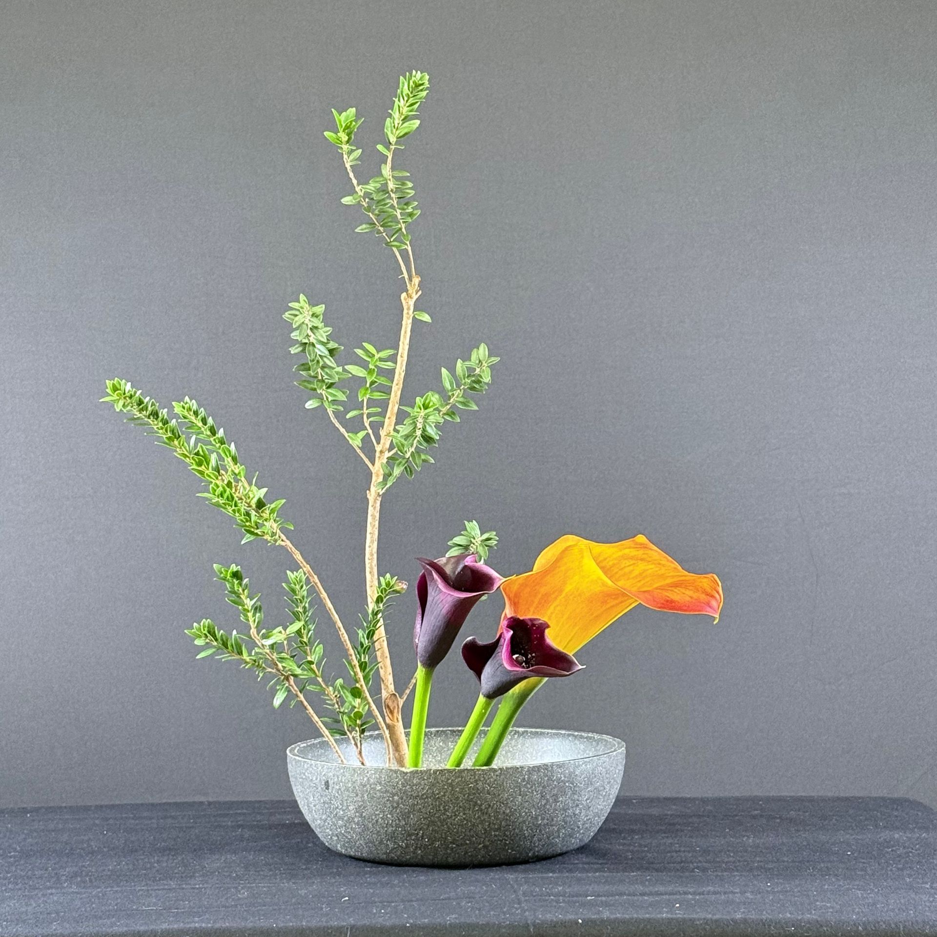 Ikebana bloemstuk: Paarse calla lelies en oranje klaprozen in een grijze stenen schaal, met bladerrijke takken.