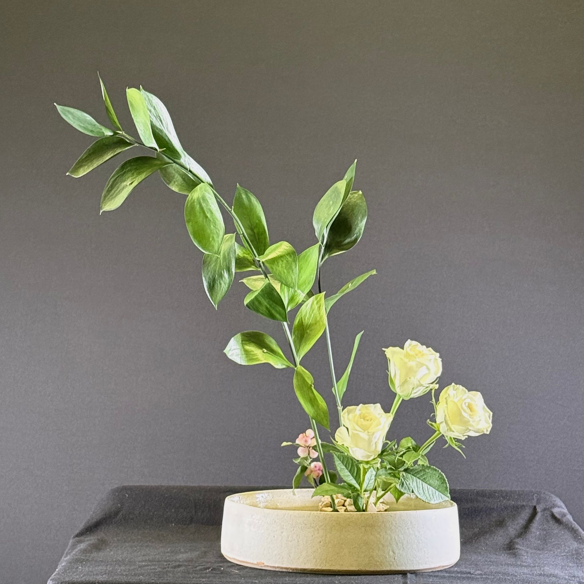 Bloemstuk: gele rozen, groen in een lage, ronde beige vaas op een donkere ondergrond.