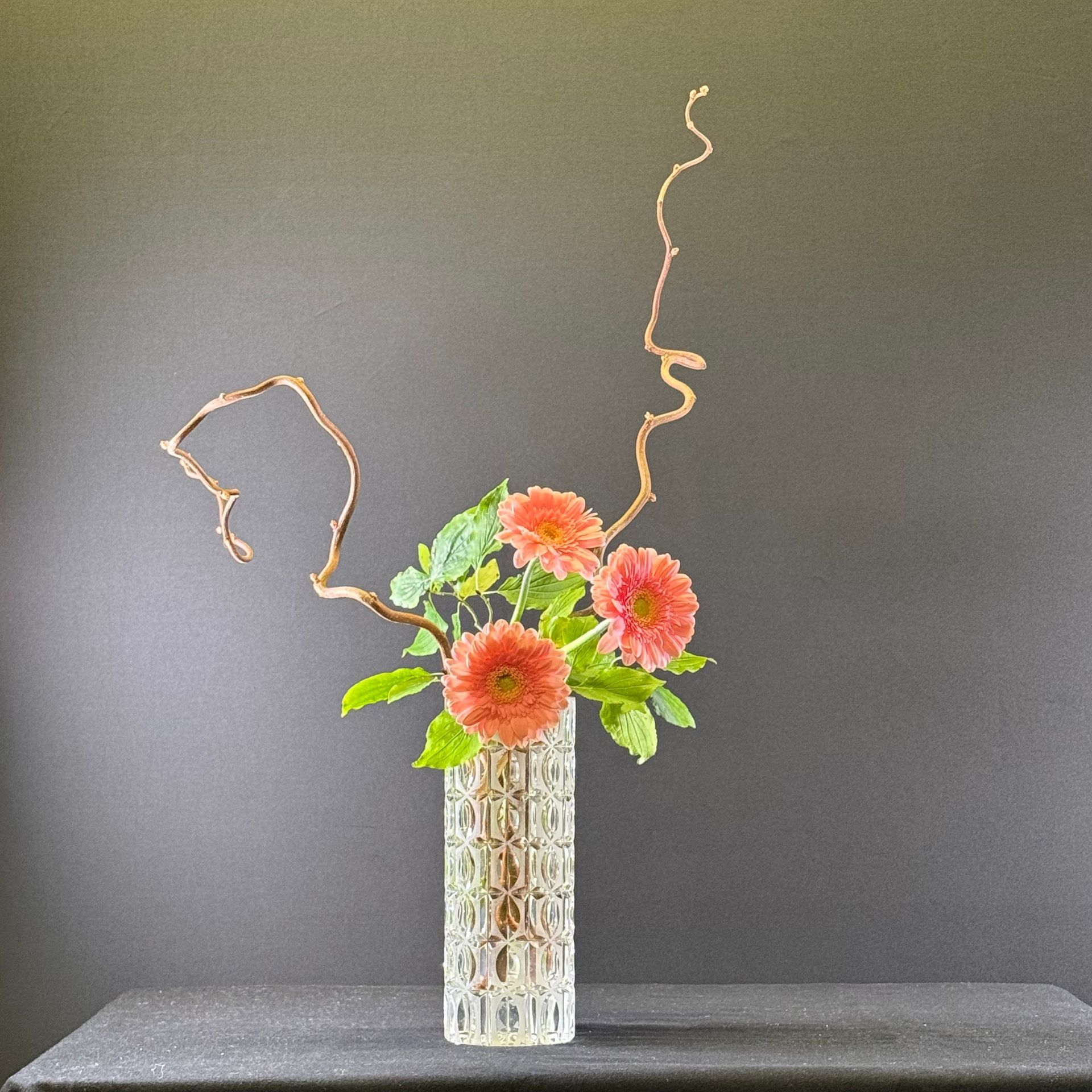 Bloemstuk in een vaas met textuur, met oranje bloemen en takken tegen een donkere achtergrond.