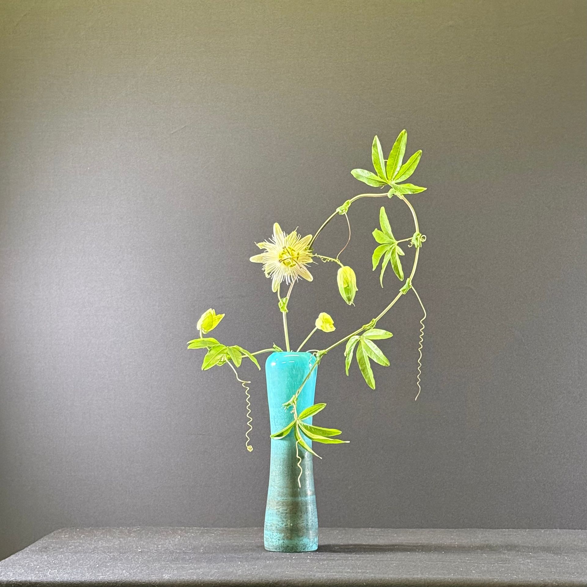 Bloemstuk in een blauwe vaas, met gele bloemen en groene bladeren tegen een grijze achtergrond.