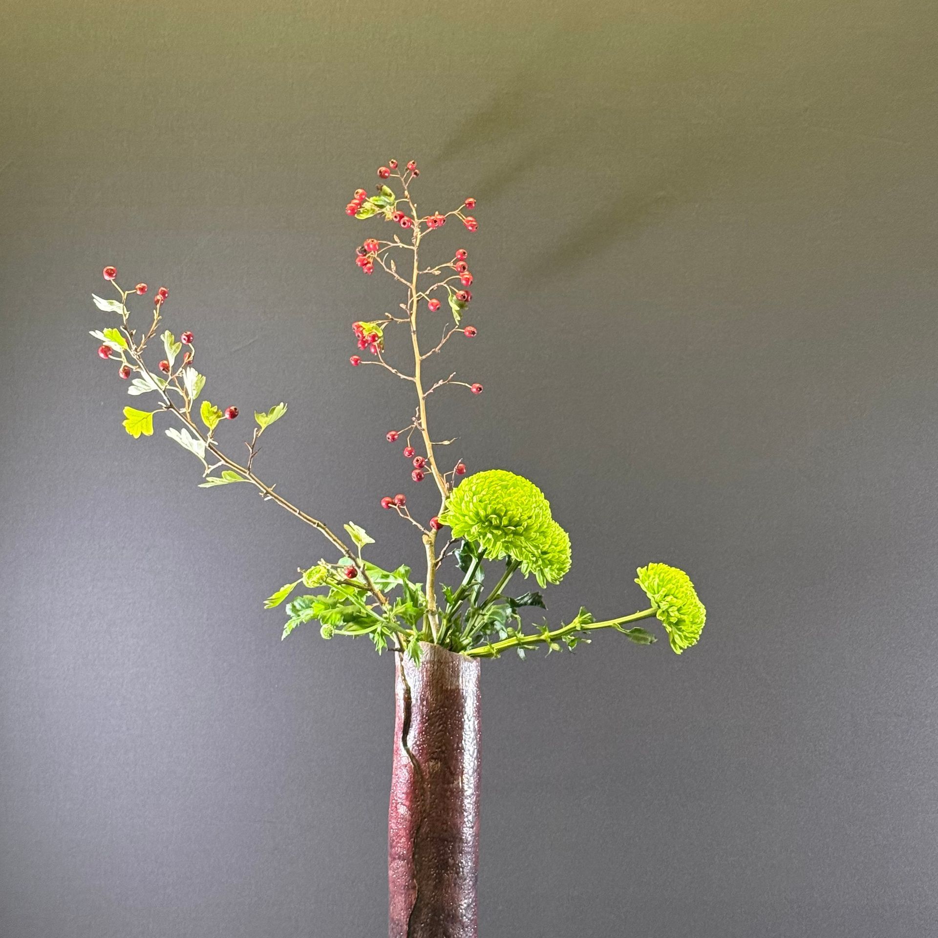 Bloemstuk met rode bessen, groene chrysanten en bladerrijke stengels in een hoge, bruine vaas tegen een grijze achtergrond.