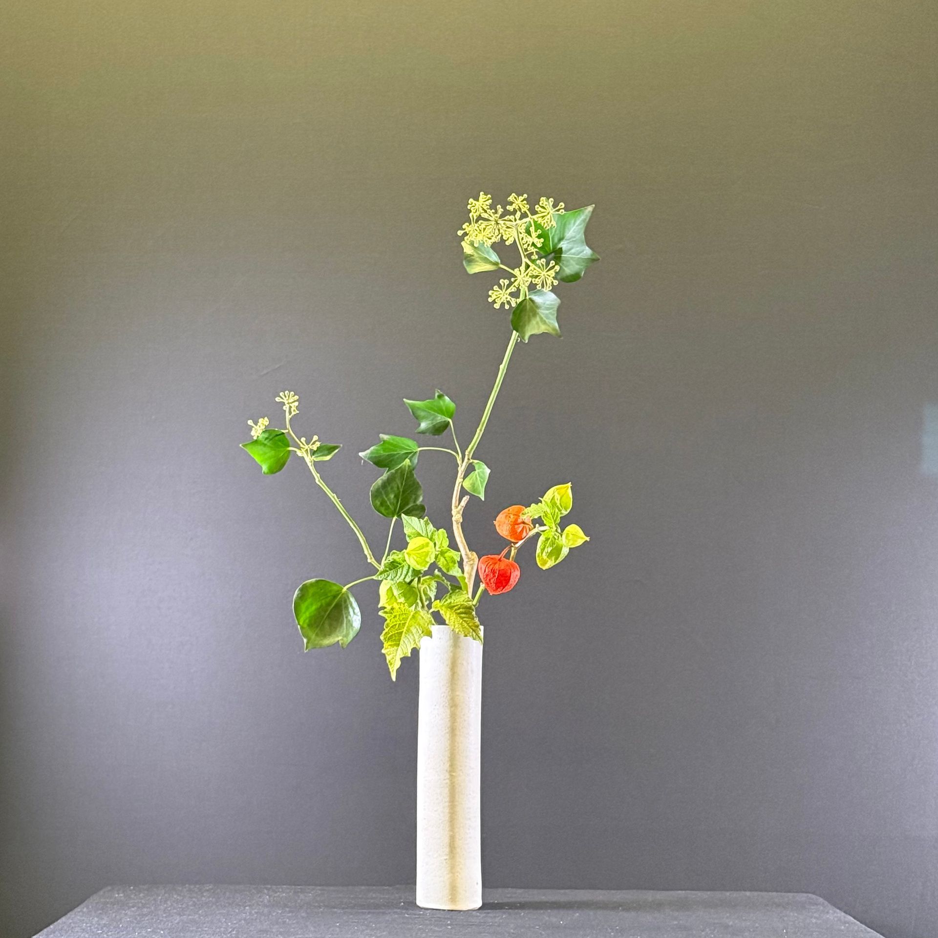 Bloemstuk in een cilindrische, gebroken witte vaas tegen een grijze achtergrond. Het arrangement bestaat uit groene stelen en kleine gele bloemen.