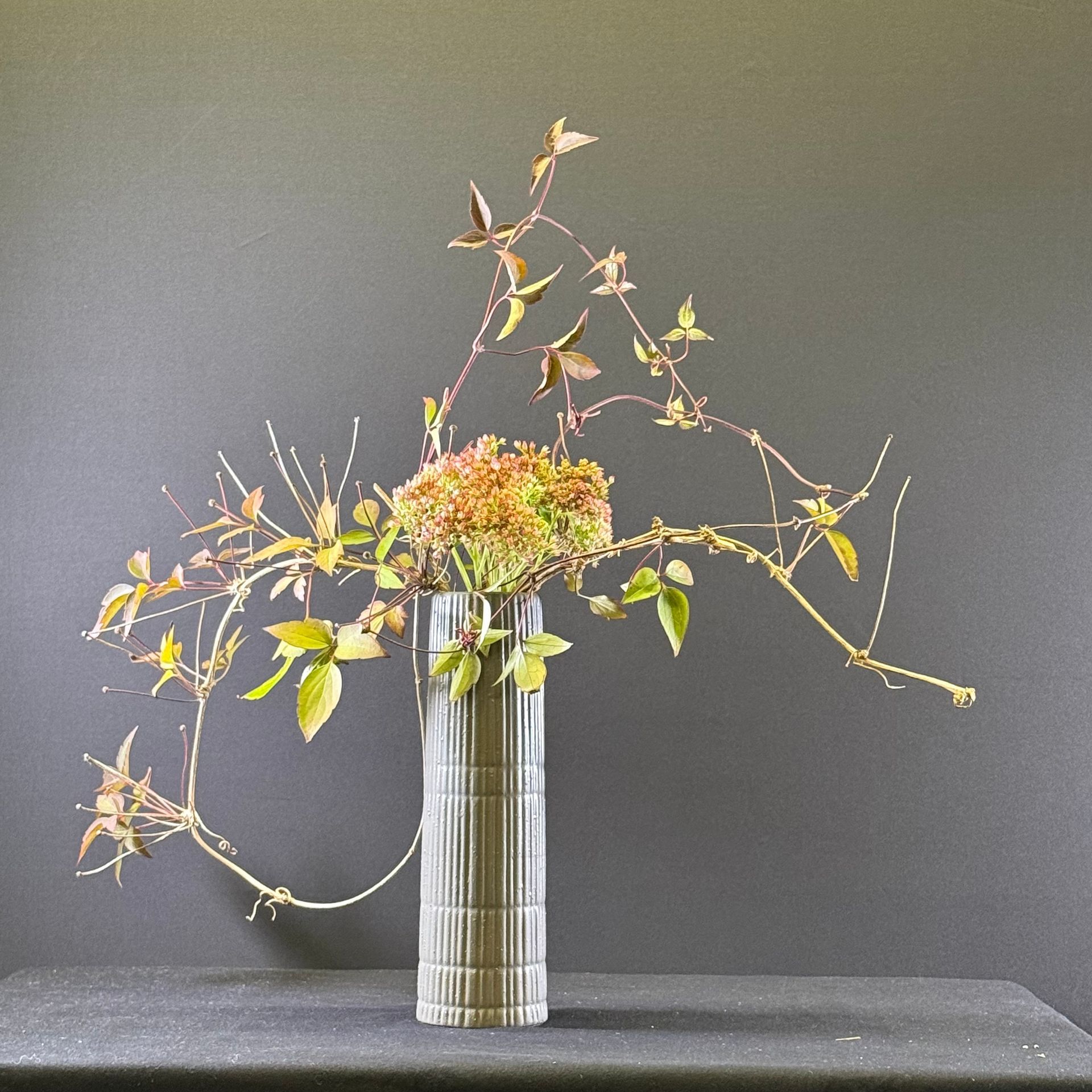 Bloemstuk in een hoge glazen vaas met klimplanten en groen op een donkere achtergrond.