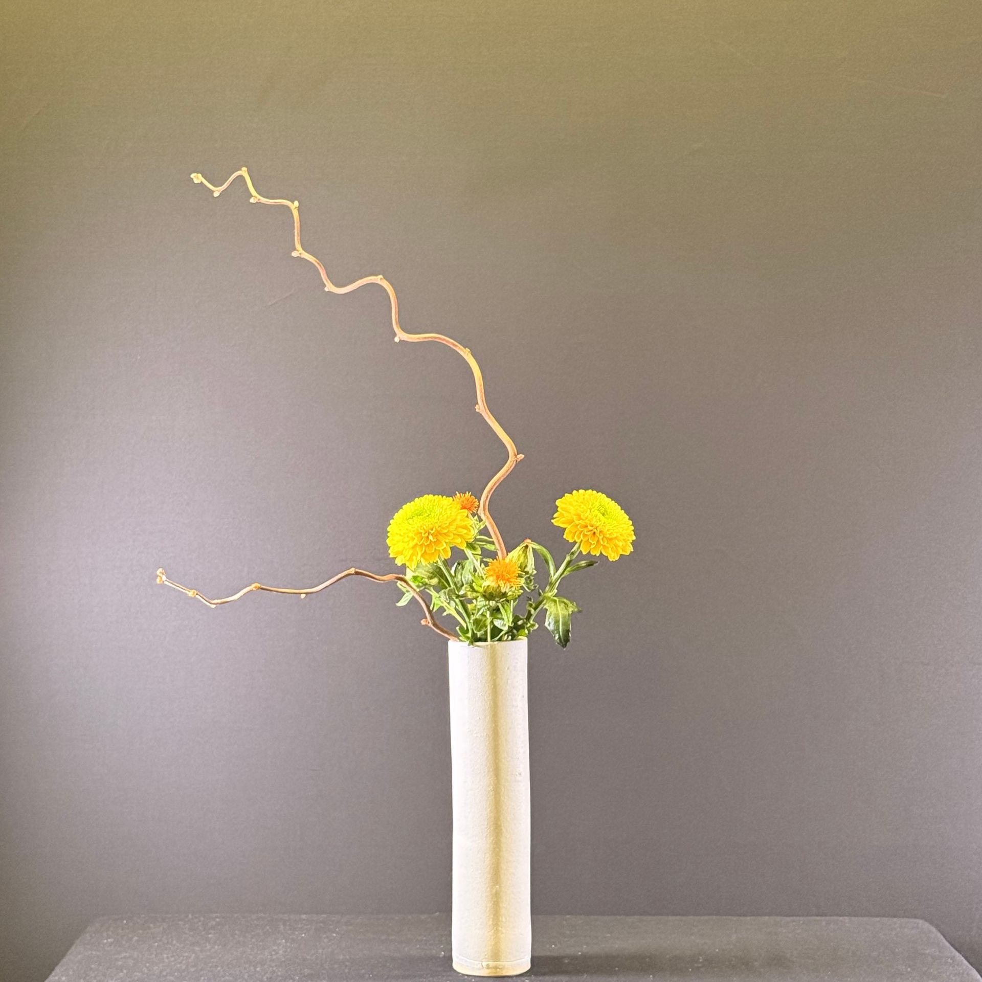 Gele bloemen en een tak in een crèmekleurige vaas tegen een grijze achtergrond.