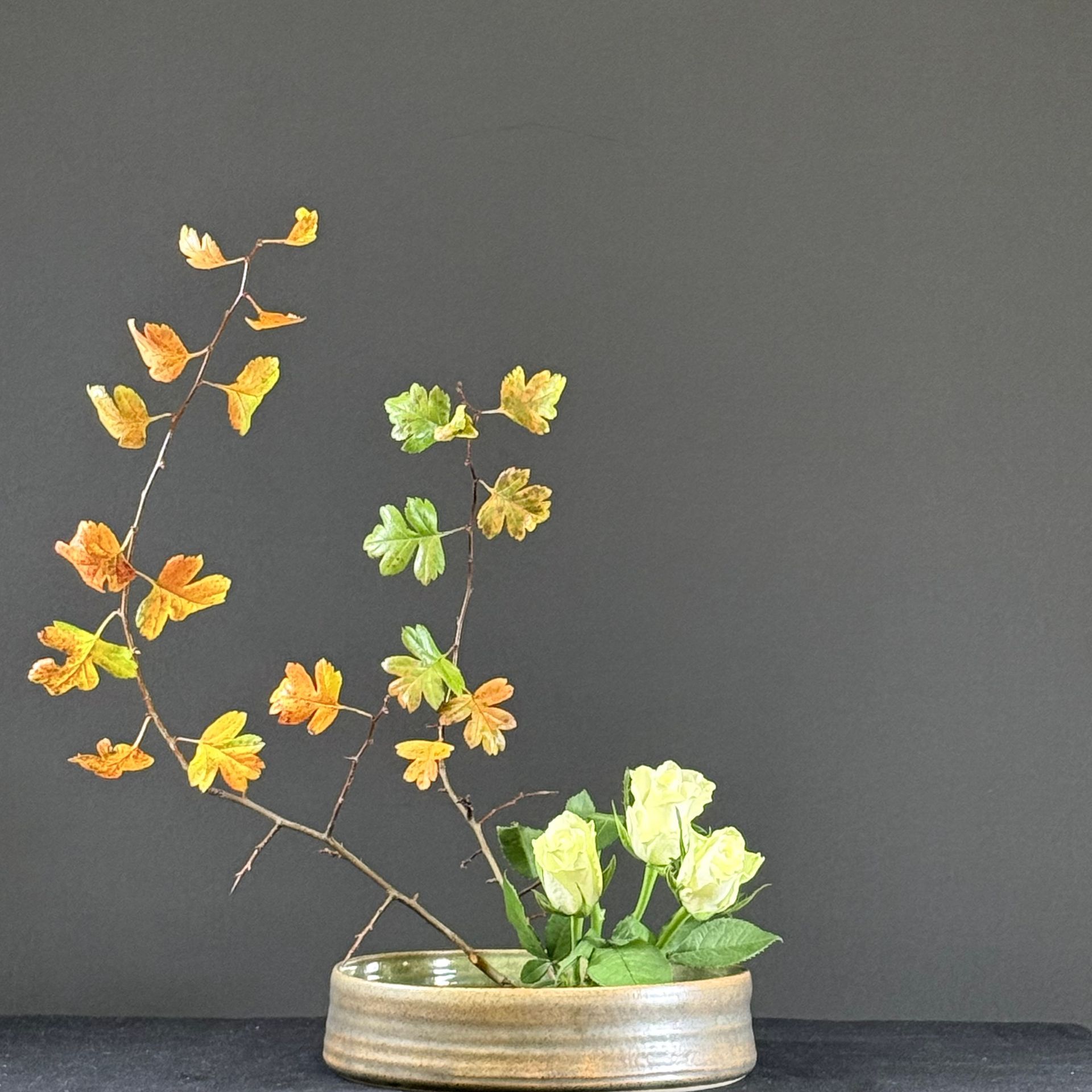 Bloemstuk met gele en groene bladeren en crèmekleurige bloemen in een lage, bruine keramische vaas tegen een grijze achtergrond.
