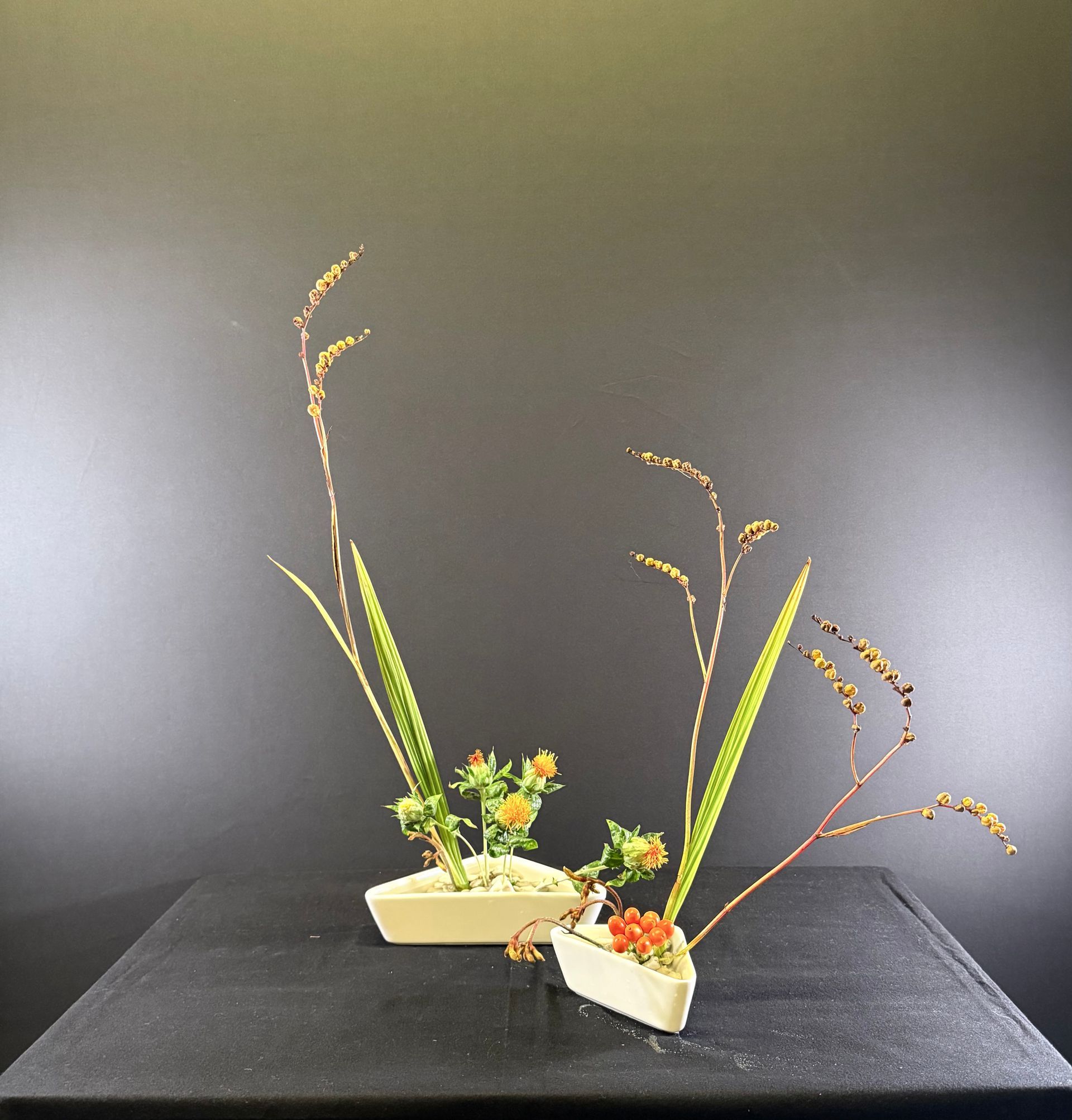 Bloemstuk met hoog gras, oranje bloemen en groene bladeren in twee witte rechthoekige vazen ​​op een zwarte achtergrond.