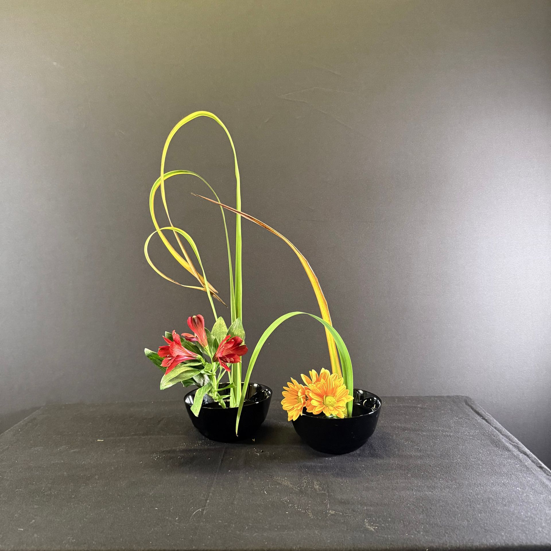 Twee zwarte schalen met bloemen en gebogen stelen tegen een donkere achtergrond. Rode en oranje bloemen.