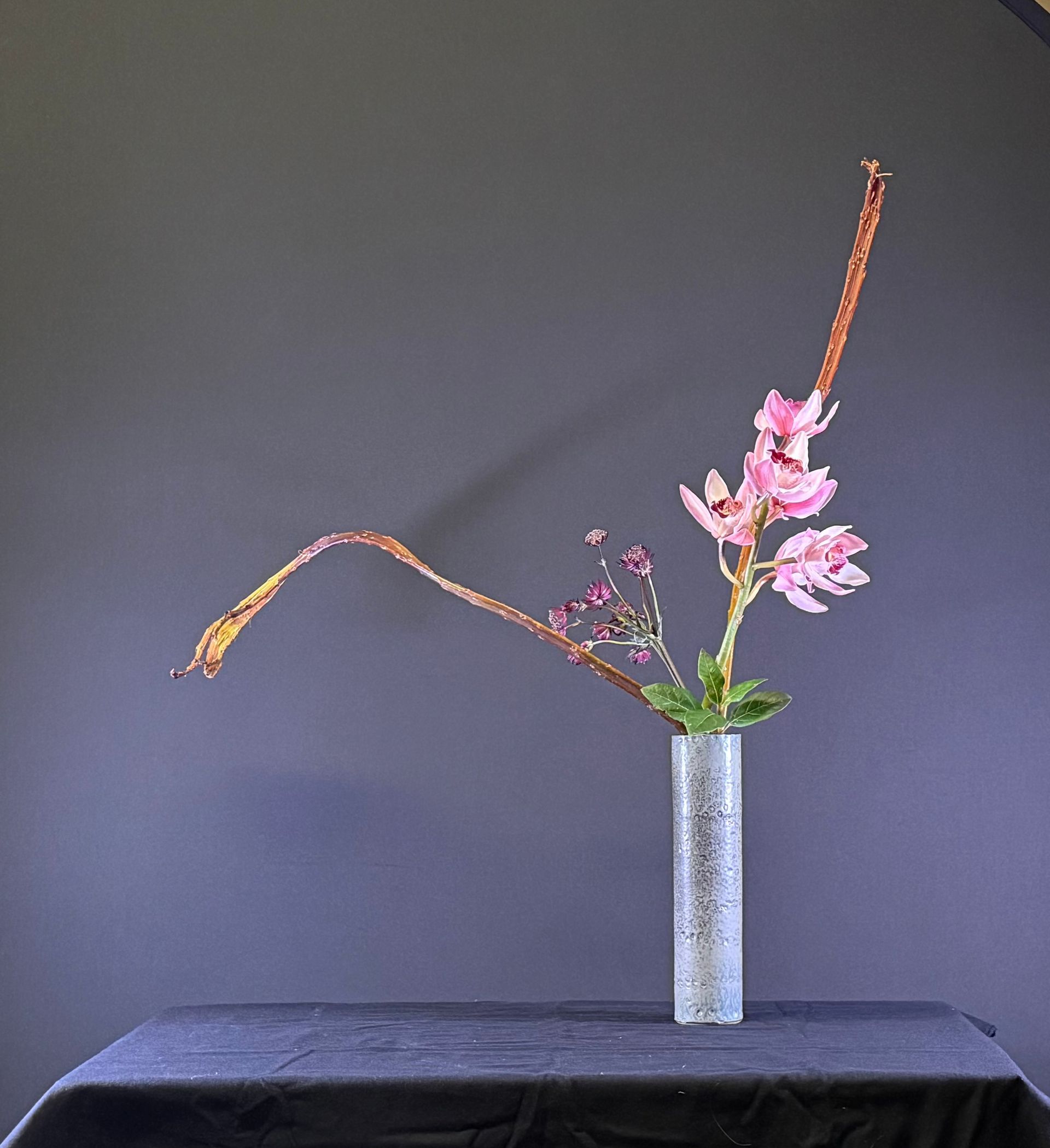 Bloemstuk in een cilindervormige vaas. Roze bloemen en slanke bruine stelen tegen een donkere achtergrond.