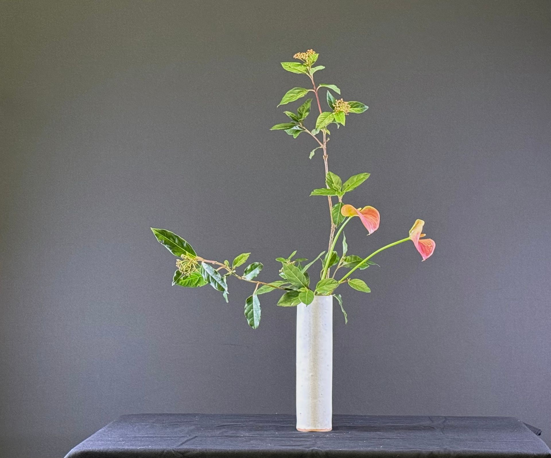 Japans bloemstuk: groene en roze takken met bladeren in een witte cilindervormige vaas op een zwarte tafel.