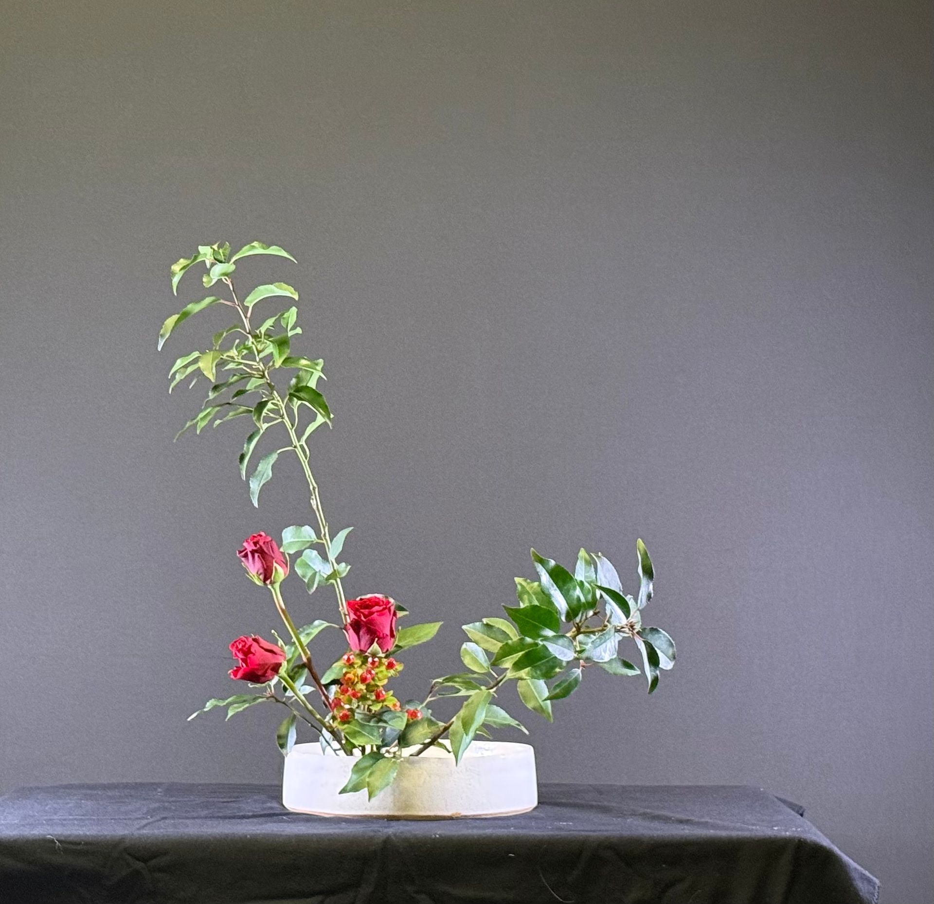 Bloemstuk met rode rozen en groen in een witte vaas op een zwarte ondergrond tegen een grijze achtergrond.