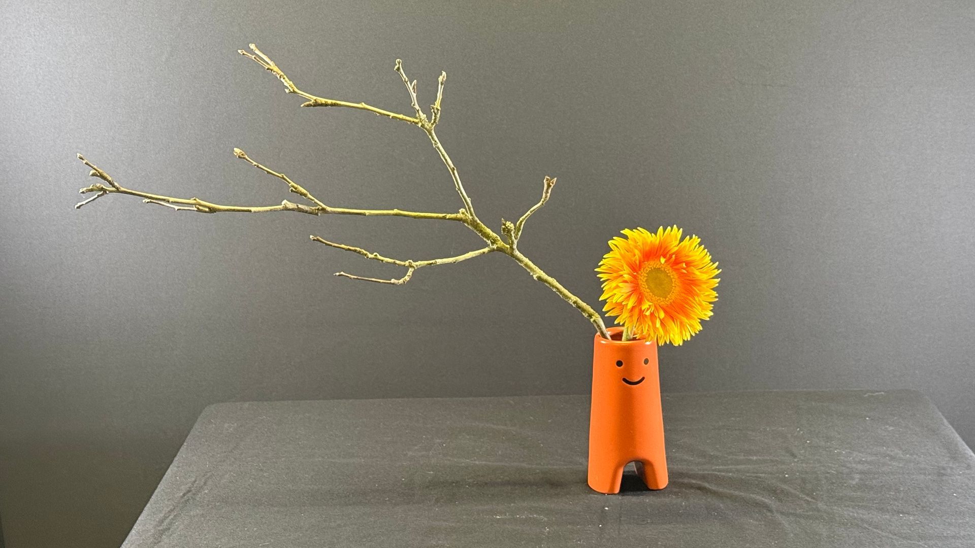 Een oranje vaas met een gele bloem en gouden takken op een tafel.