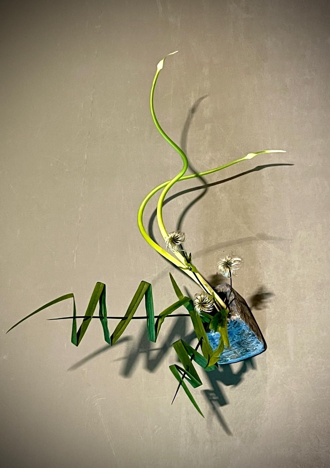Abstracte bloemschikking met groene stelen, witte knoppen en blauwe steen op een grijze achtergrond.