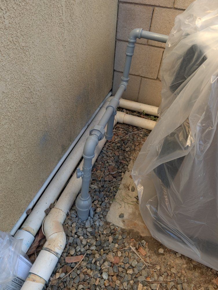 Outdoor plumbing pipes and PVC lines beside a stucco wall, next to gravel and a covered object.