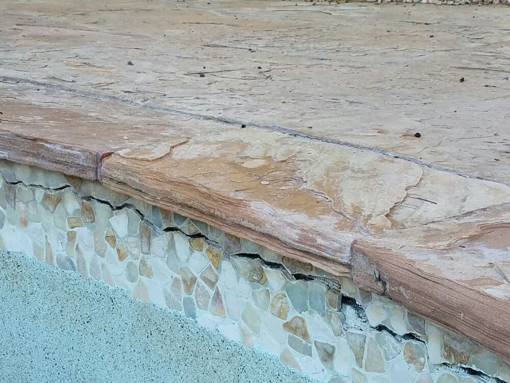 Stone pool edge with tan coping and mosaic tile trim above blue water