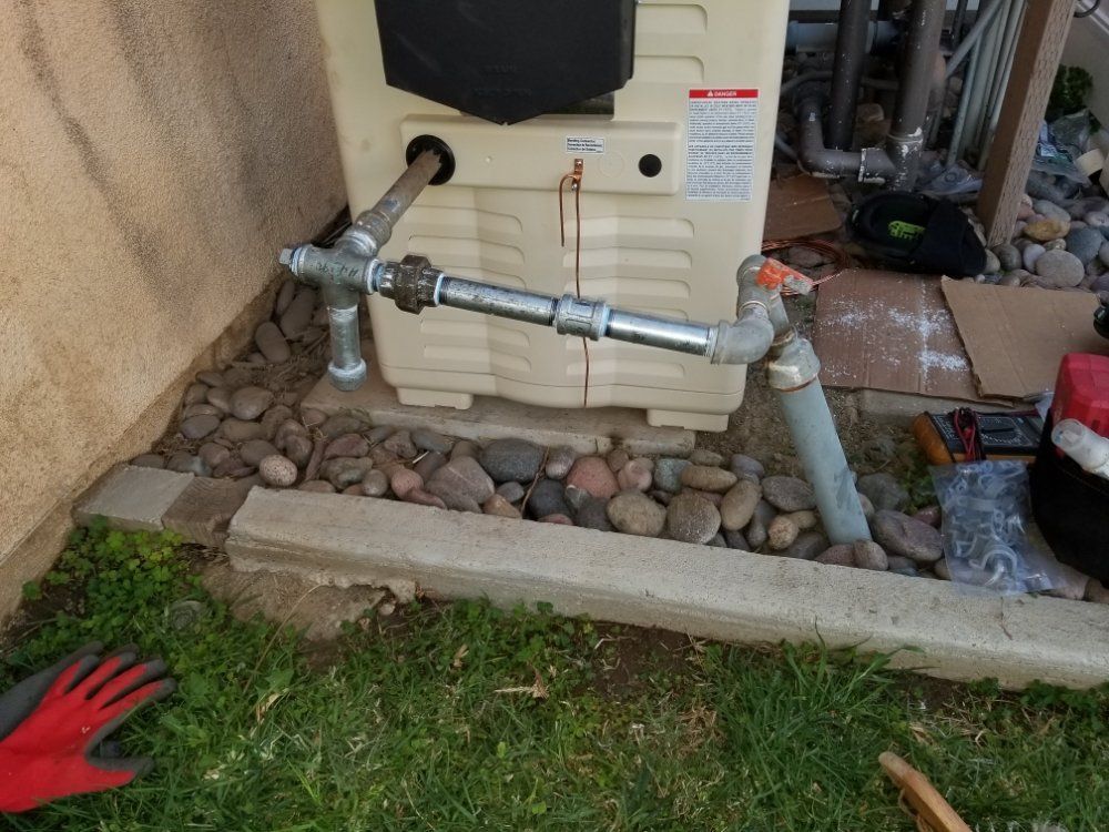 Outdoor plumbing pipes and valves beside a house wall, with rocks and grass below.