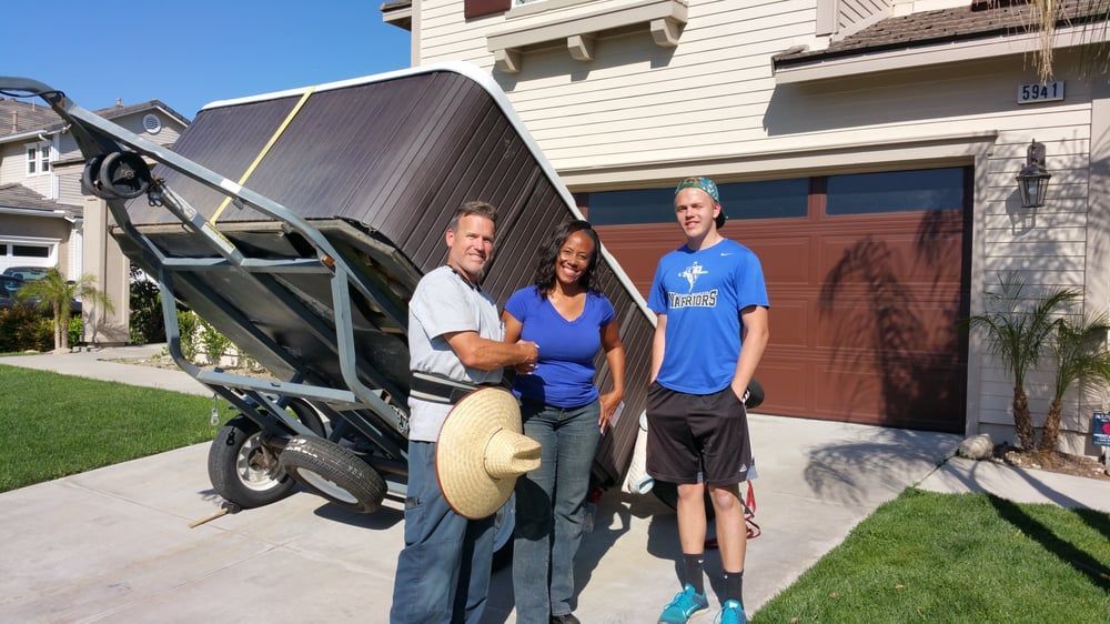 Three people standing by a driveway beside a solar panel delivery truck and a suburban house.