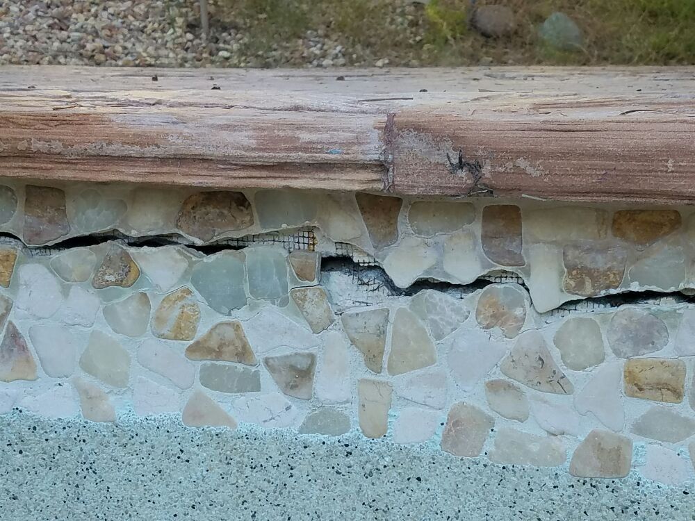 Stone retaining wall with cracked mortar beneath a wooden beam, showing visible damage.