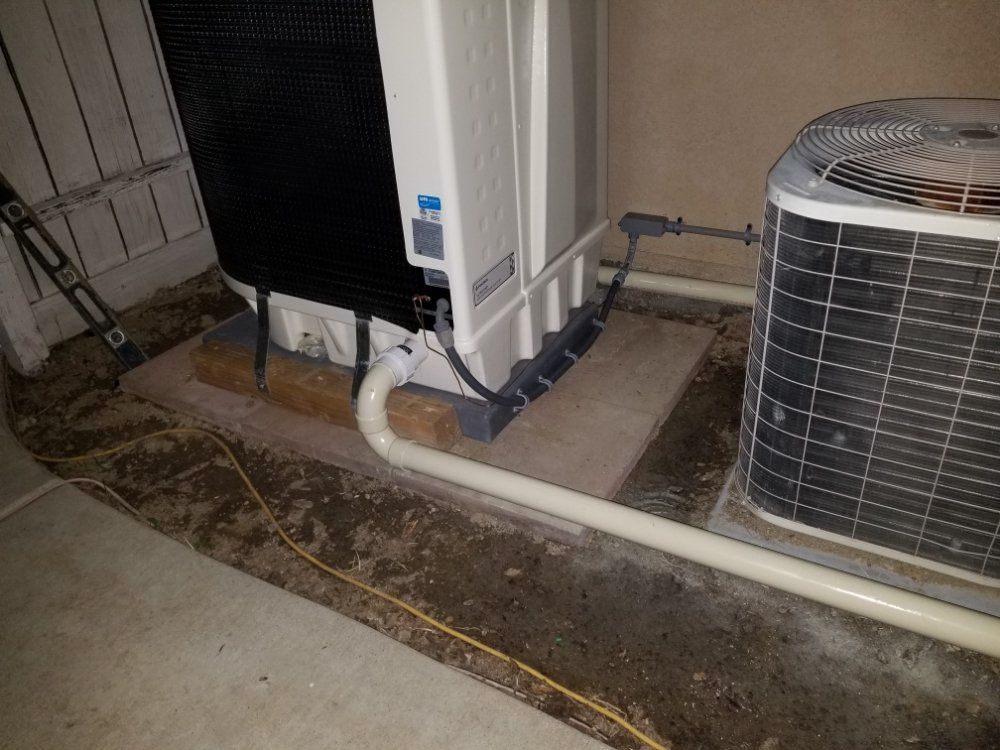 Outdoor HVAC unit beside a building with white piping and a black utility box on a concrete pad