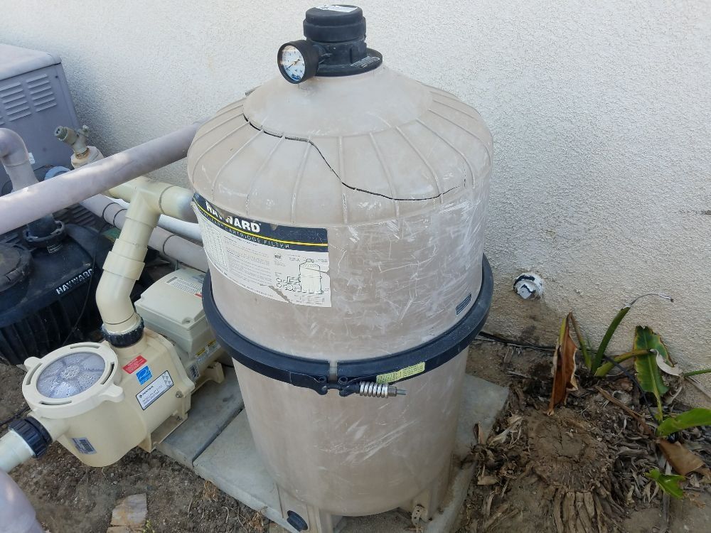 Pool filter tank and pump equipment beside a wall, with a pressure gauge on top.