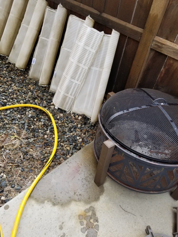 Backyard corner with stacked rolled mats, a metal fire pit, and a yellow hose on wet concrete.