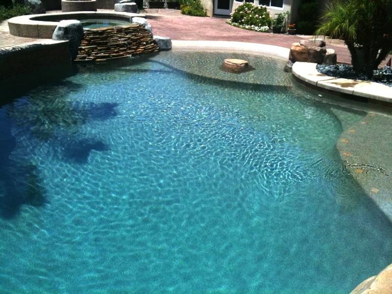 Blue swimming pool with stone waterfall and surrounding patio in a sunny backyard