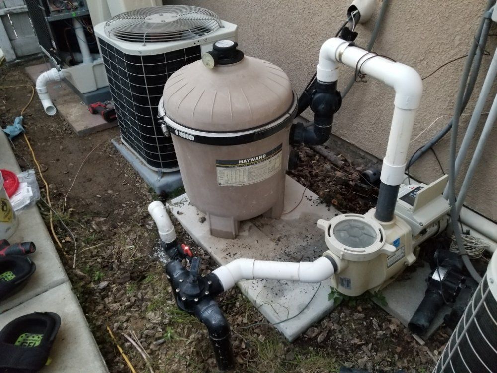 Outdoor pool equipment: beige filter tank, white pipes, pump, and HVAC unit beside a house.