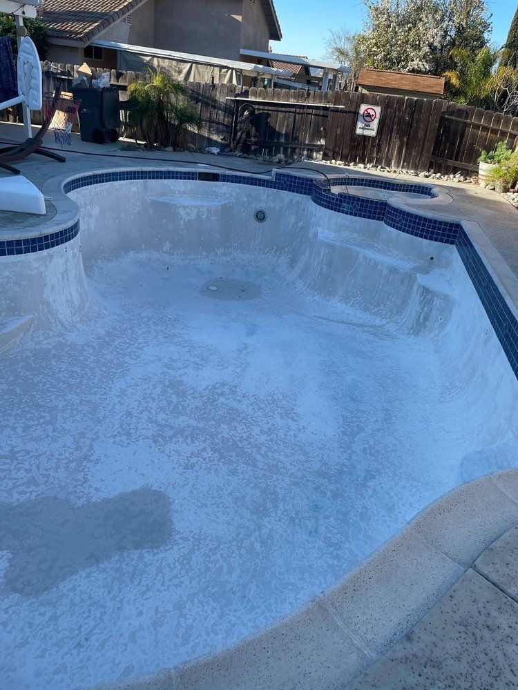 Empty swimming pool with blue tile trim and a cracked, peeling interior beside a backyard fence.