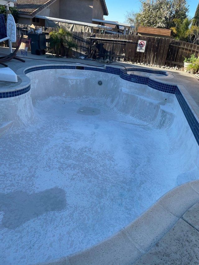 Empty swimming pool with blue tile trim and a cracked, peeling interior beside a backyard fence.