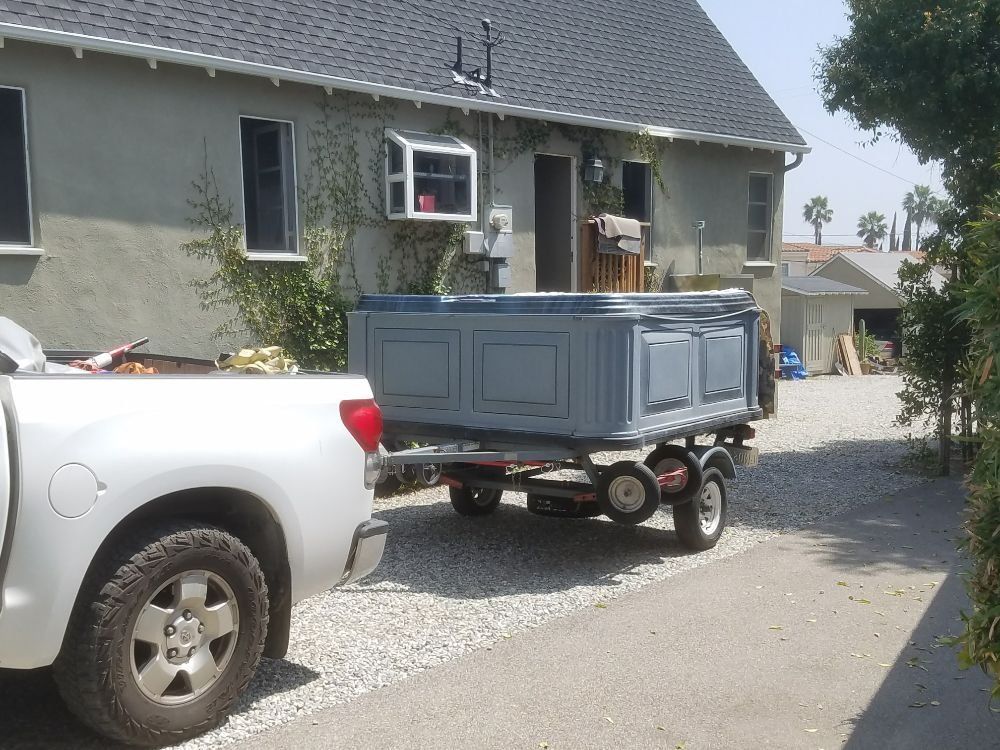 Gray pickup towing a blue utility trailer in a gravel driveway beside a house.