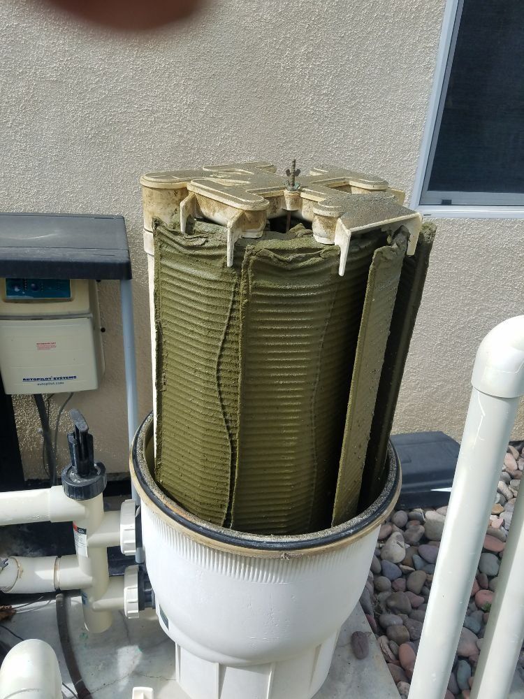 Open pool filter cartridge with green pleated elements in a white housing next to pipes and rocks.