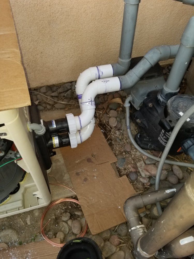 Plumbing pipes and equipment in a cramped utility area, with white and gray PVC pipes over cardboard on the floor.