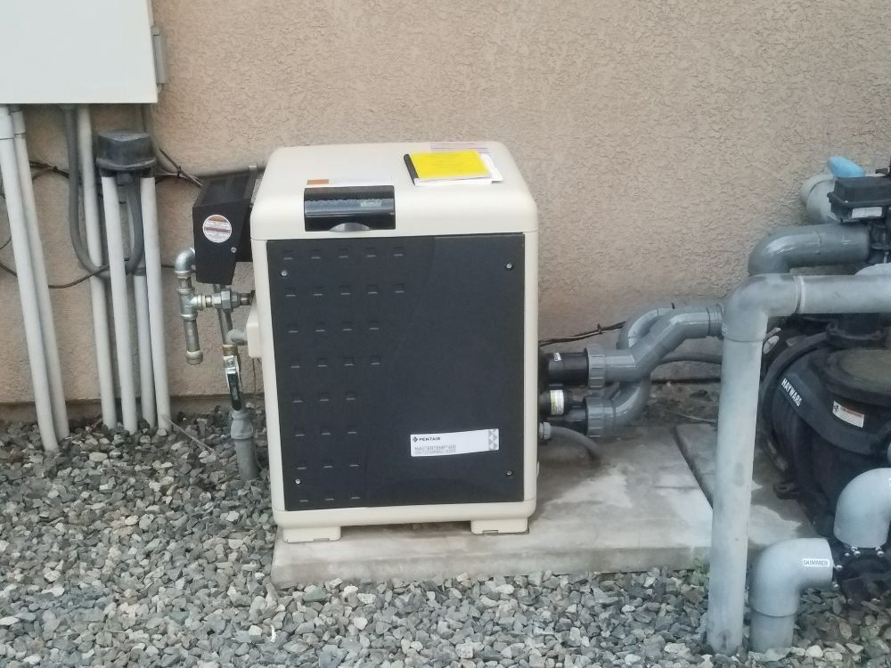 Outdoor HVAC unit beside a house wall, with pipes and gravel around it