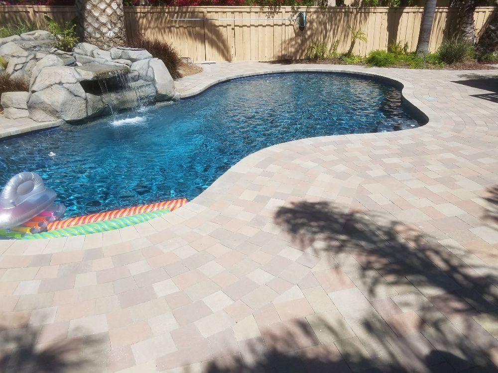 Backyard swimming pool with blue water, stone waterfall, and light paver patio in sunlight