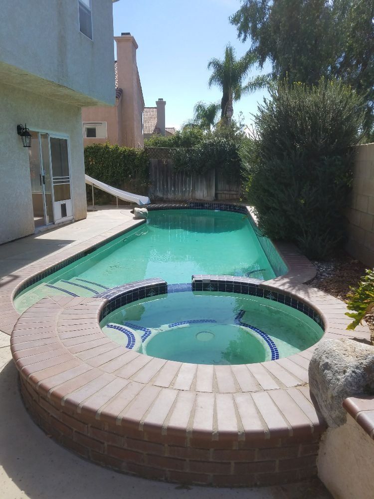 Backyard pool with a small hot tub and turquoise water beside a house and tall shrubs