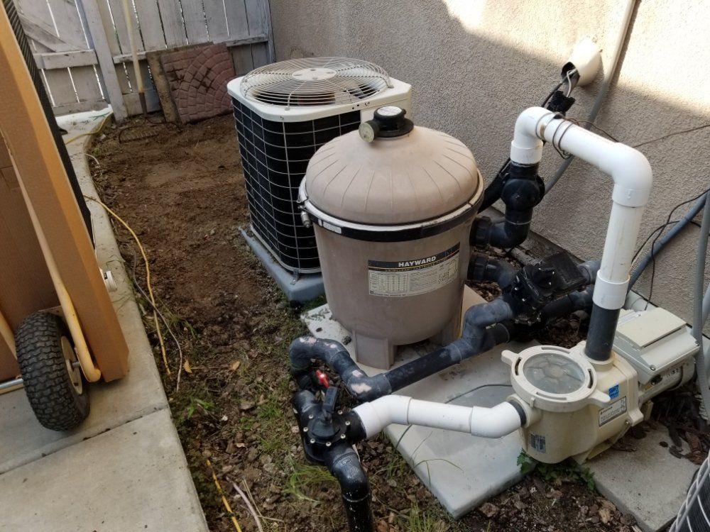 Pool pump and filter equipment with pipes beside a house in a narrow outdoor yard.