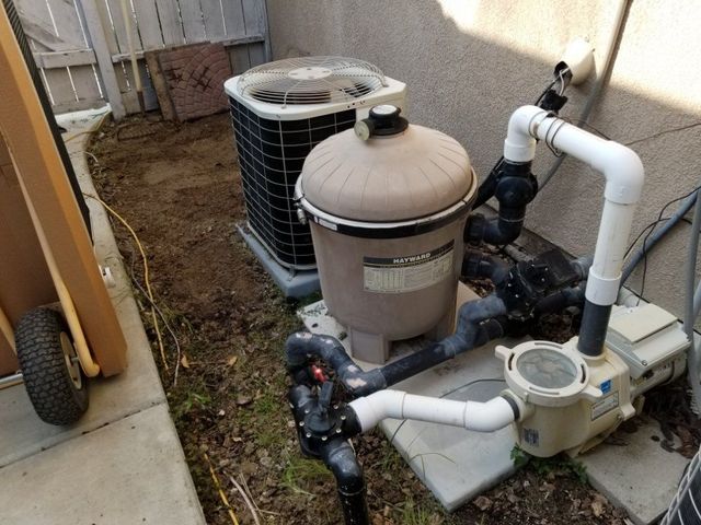 Pool pump and filtration equipment with white pipes and tan tank beside a house in a narrow outdoor space
