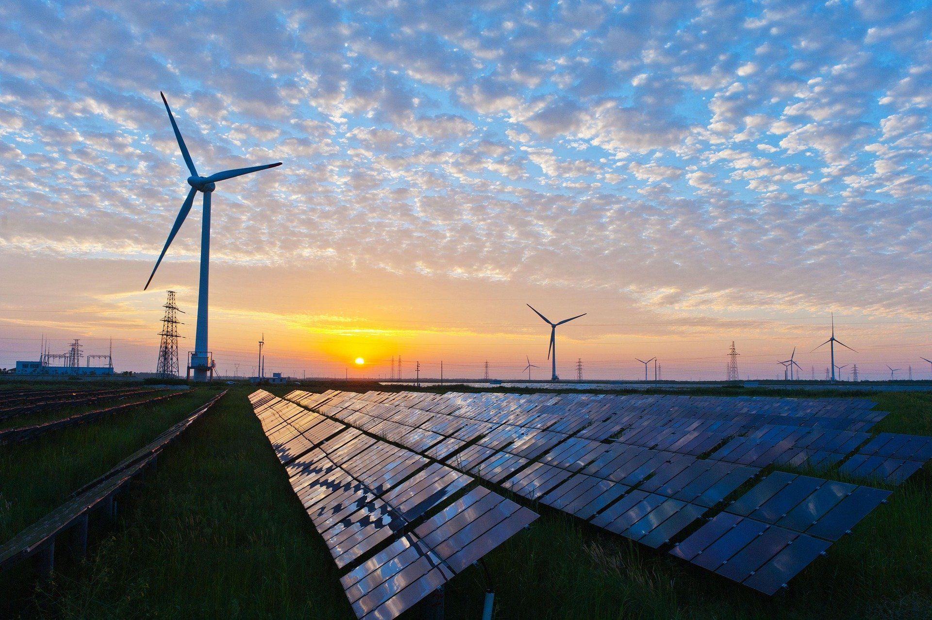 A field of solar panels and wind turbines at sunset.