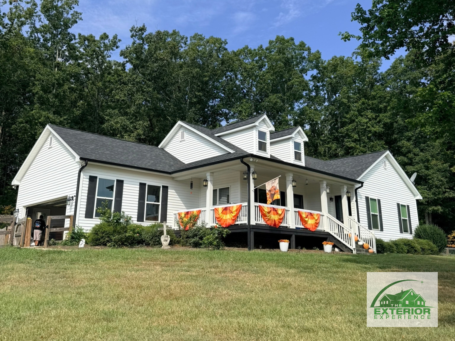 White house with black shutters and colorful porch flags on a grassy hill, backed by trees