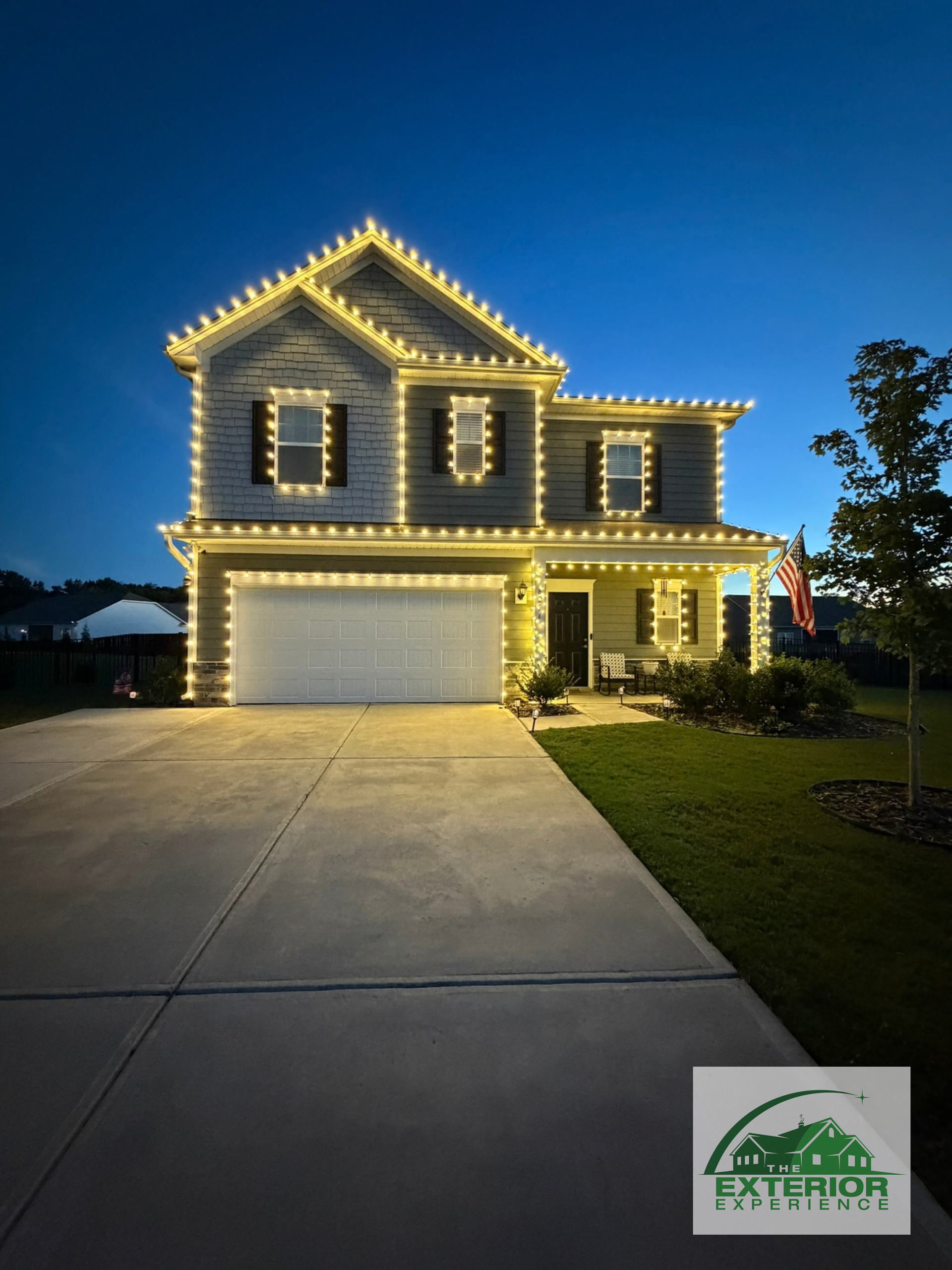 Two-story house outlined in white lights at dusk, with a wide driveway and manicured lawn.