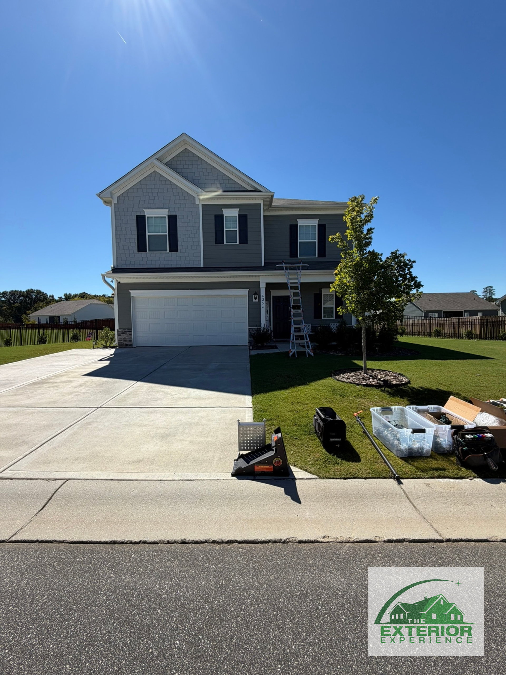 Two-story gray house with white garage and a driveway under a clear blue sky, with landscaping supplies in front.