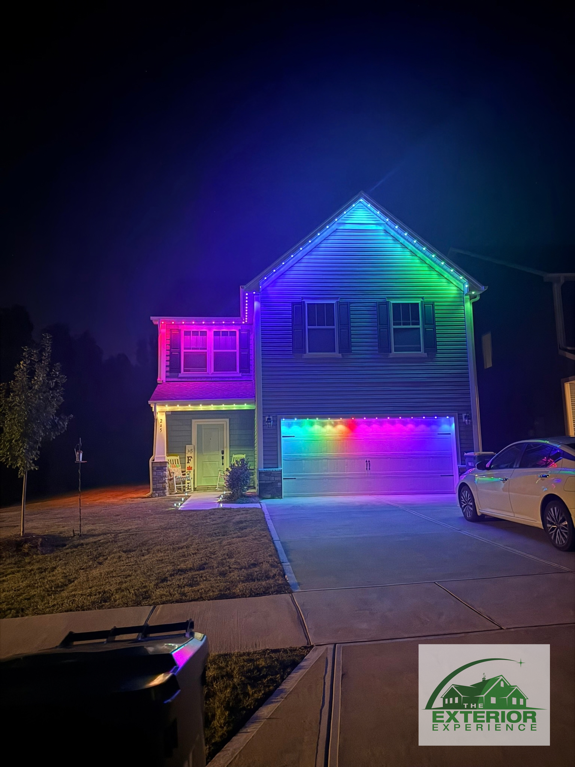 Two-story house lit with rainbow holiday lights at night, with a car parked in the driveway.