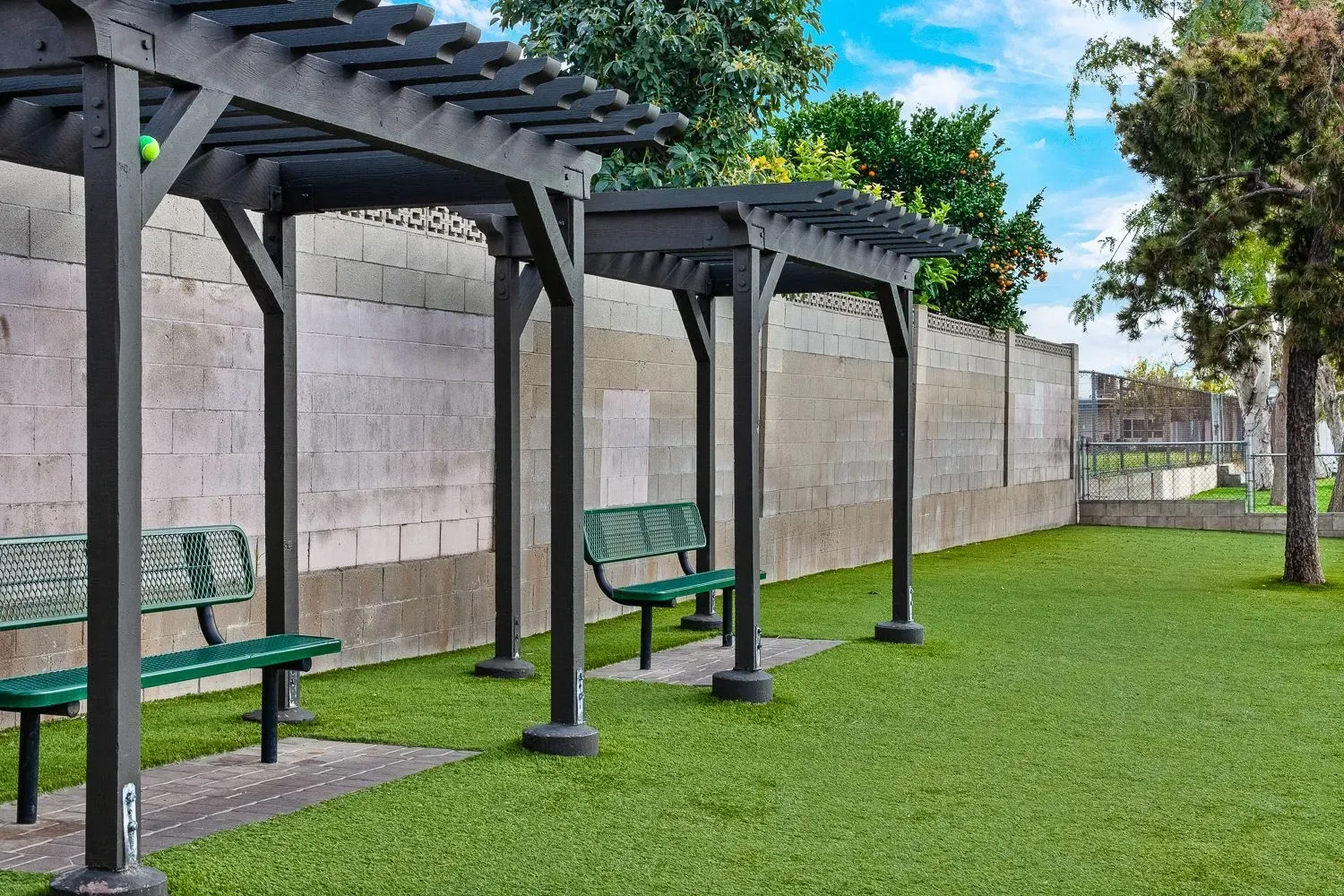 Park with green benches under black pergolas beside a stone wall and grassy lawn.
