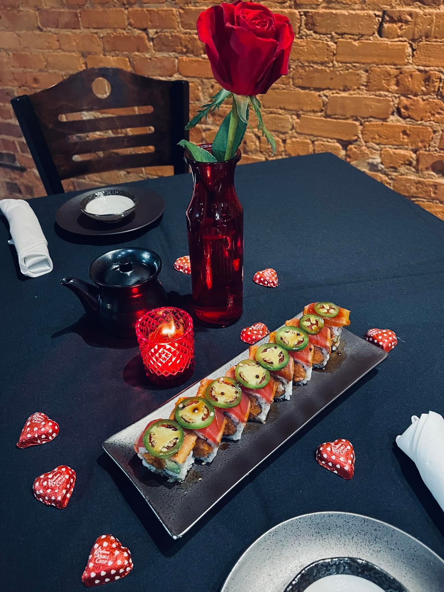 Sushi dinner setting: red rose, candles, heart confetti, and a sushi roll on a black table.