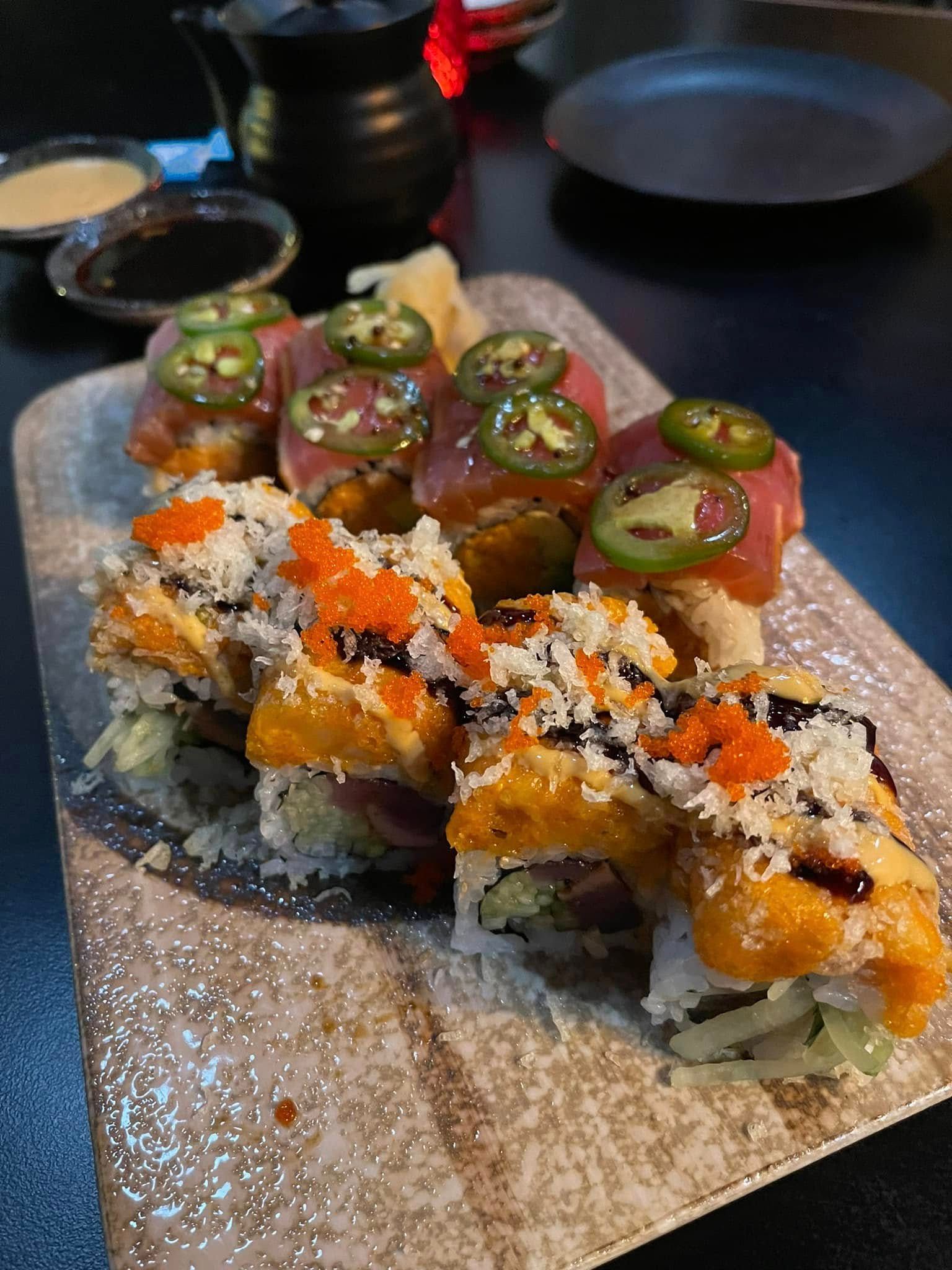 Sushi rolls with toppings of tuna and jalapeño slices, on a textured wooden board. Soy sauce and ginger are also present.