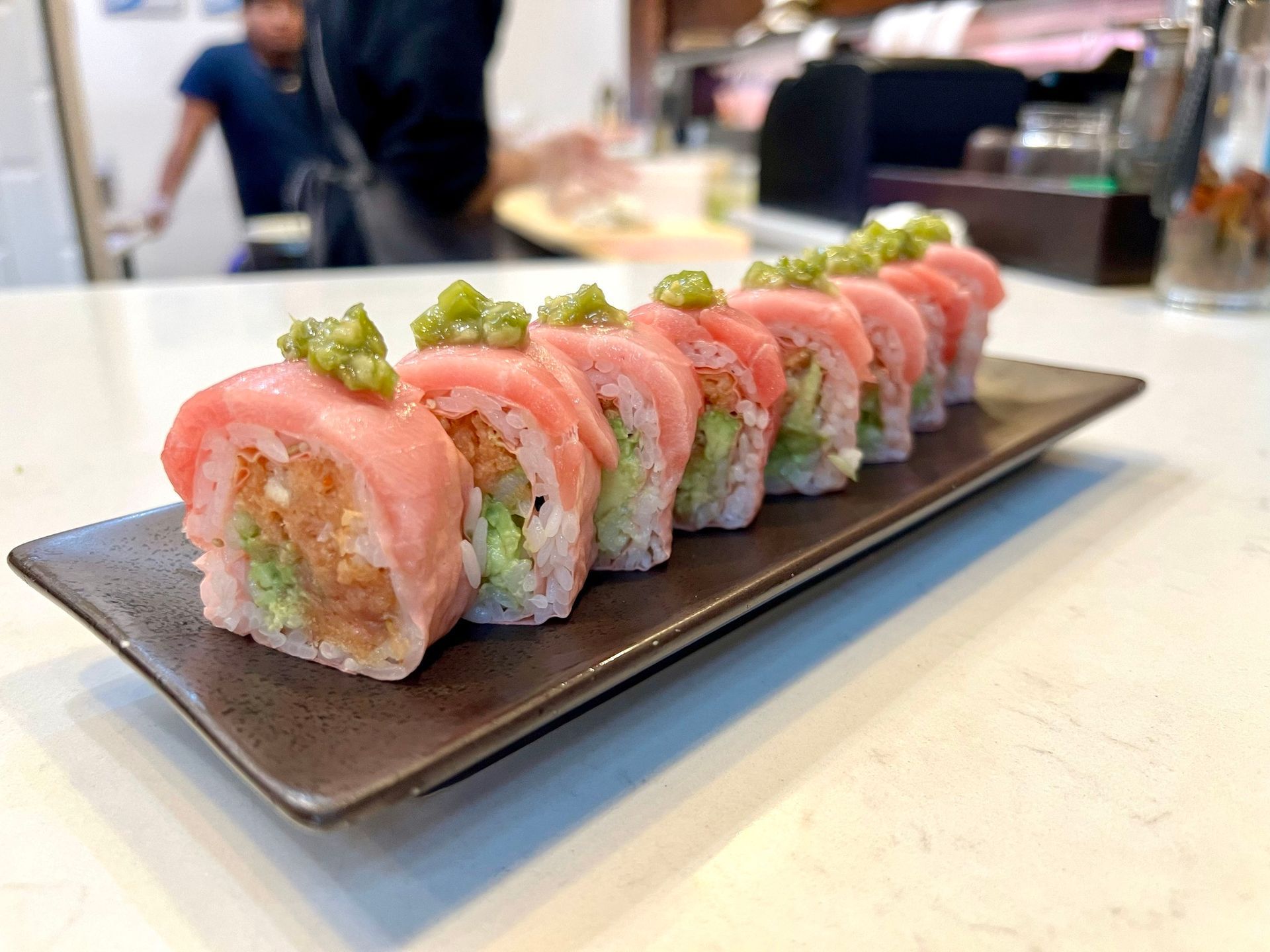 Sushi roll, pink tuna over rice with green topping, arranged on a black rectangular plate.