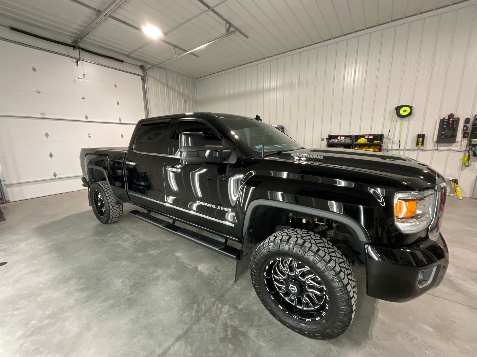 A black pickup truck with off-road tires parked inside a well-lit garage with a white metal wall and closed door.