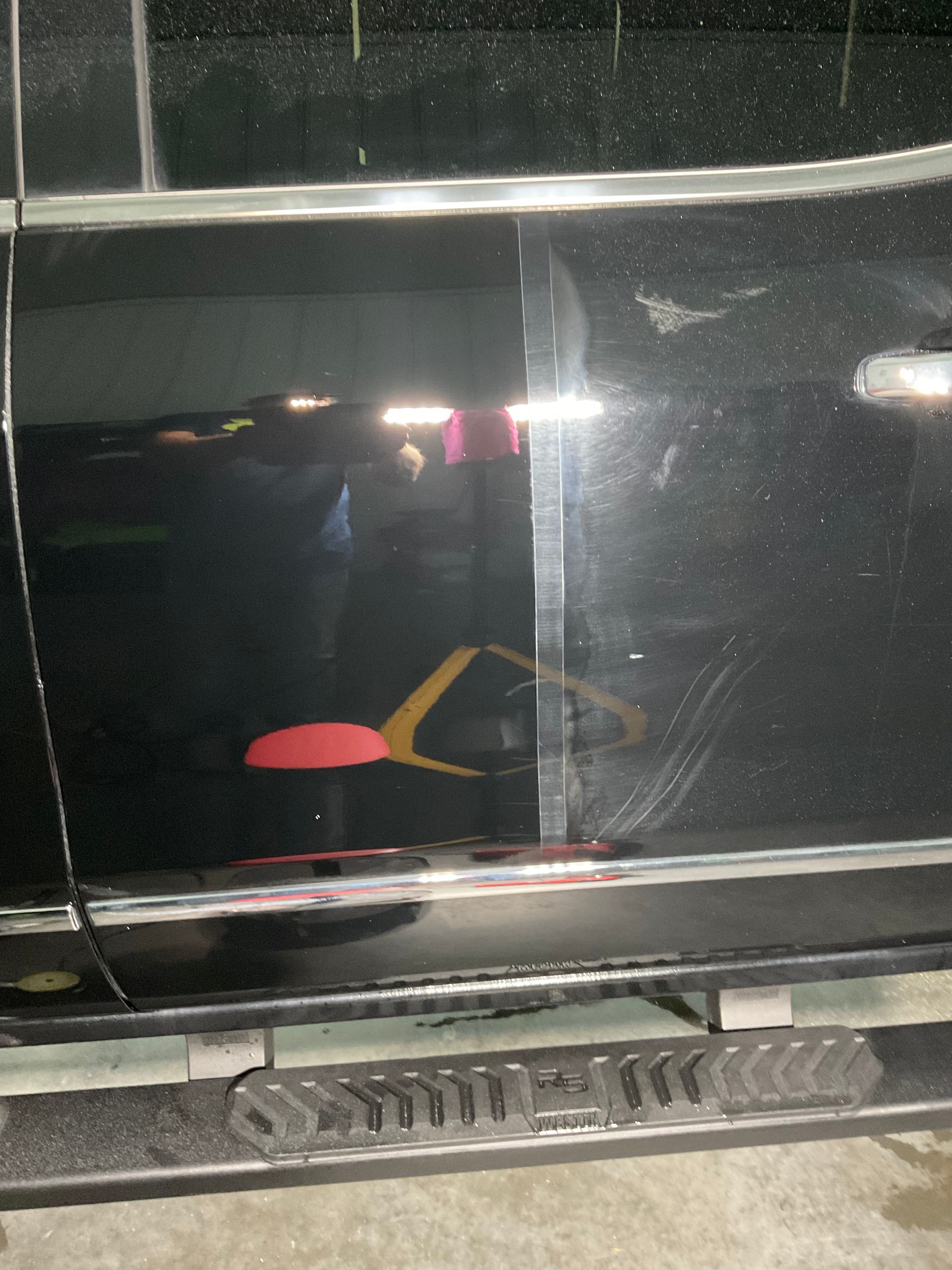 A close-up view of a black vehicle door featuring a prominent, light-colored scratch on the paint surface.