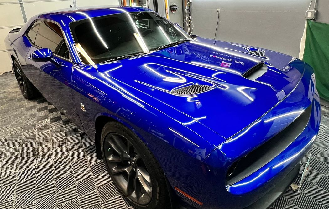 A shiny, deep blue Dodge Challenger parked inside a garage with patterned flooring and bright overhead lighting.
