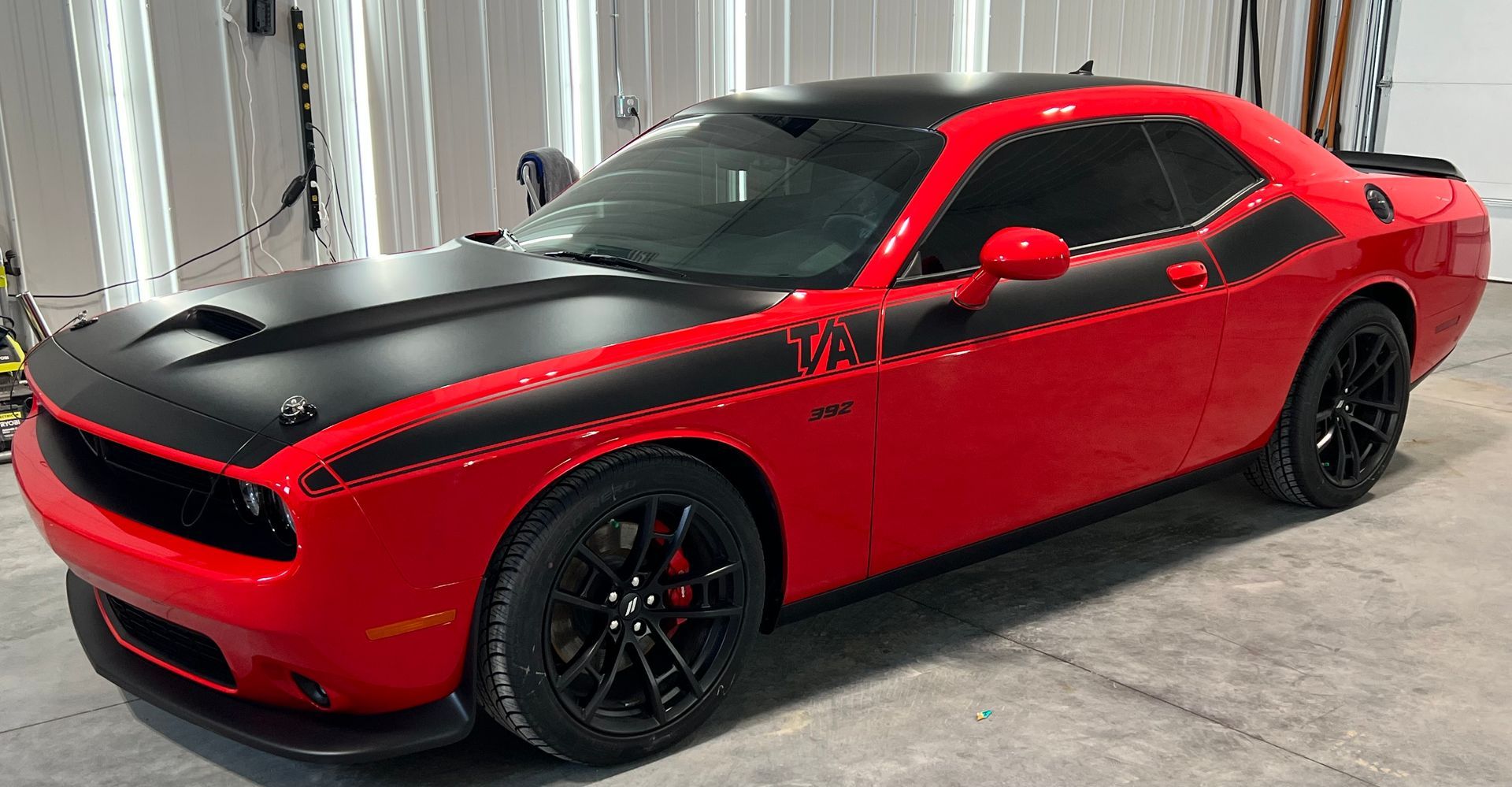 A red Dodge Challenger with a matte black hood, roof, and side stripe, parked in a well-lit garage.